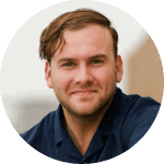 Portrait of a smiling man with short hair, wearing a navy blue shirt, set against a blurred outdoor background.