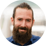 Smiling man with a beard, wearing a blue shirt, representing a professional in a modern, collaborative environment.